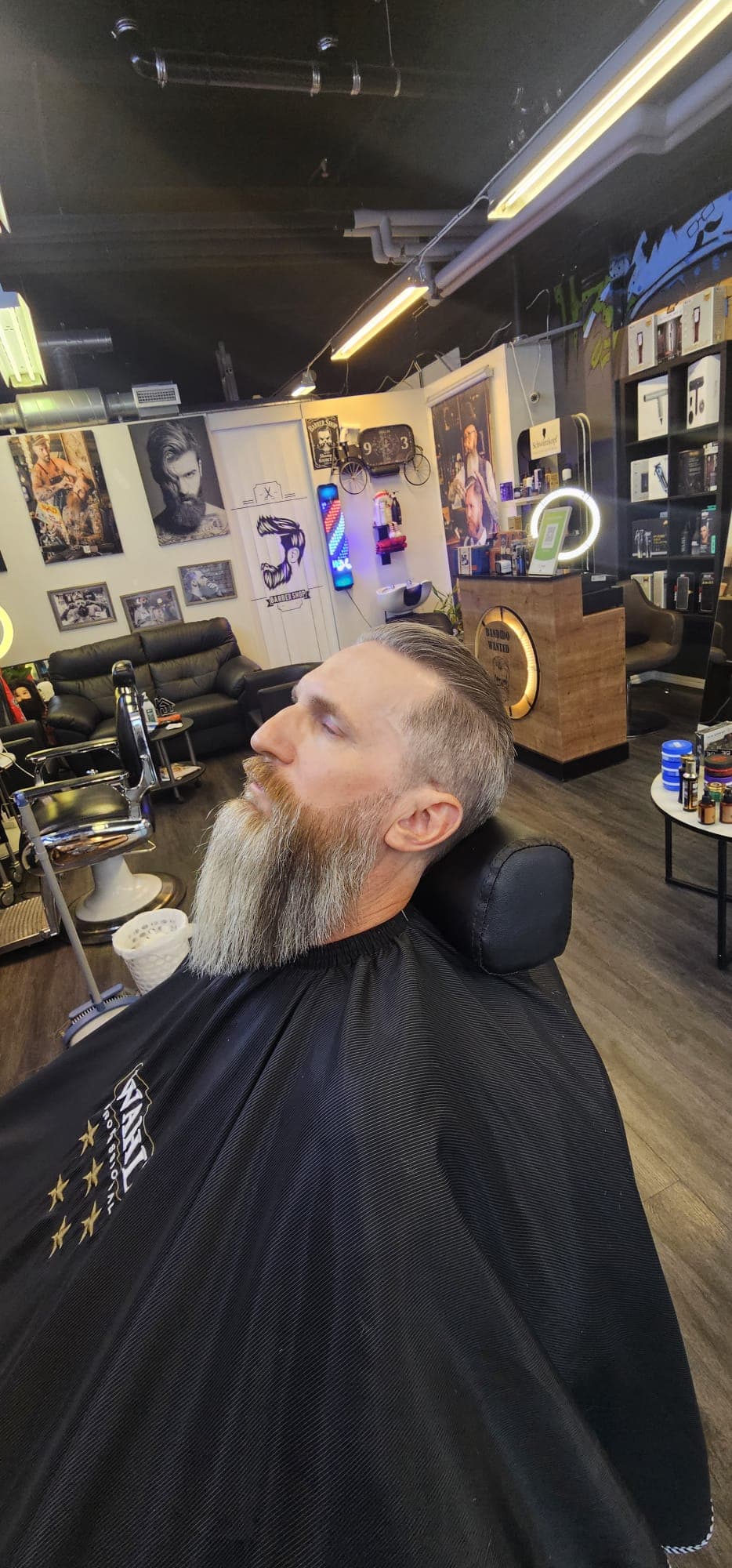 Kund med långt, välvårdat helskägg och bakåtkammat hår i barberarstolen, med salongens stolar, skinnsoffa och barberarstolpe i bakgrunden